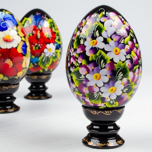 Op de afbeelding: Drie handgeschilderde houten eieren met bloemmotieven. De eieren zijn zwart met witte, paarse, rode en groene bloemen. De eieren staan op zwarte standaards.