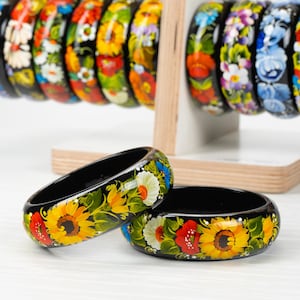 May include: Two black wooden bangles with colorful floral designs painted on them. The bangles are stacked on top of each other. The designs feature sunflowers, red poppies, and other flowers.