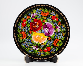 May include: A black wooden plate with a hand-painted floral design. The plate features red, yellow, purple, and blue flowers with green leaves and red berries. The plate is on a black stand.