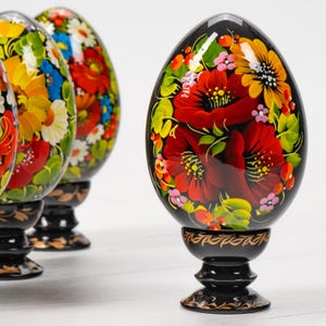 May include: Five black wooden eggs with colourful floral designs painted on them. The eggs are on black wooden stands. The eggs are a traditional Russian folk art.