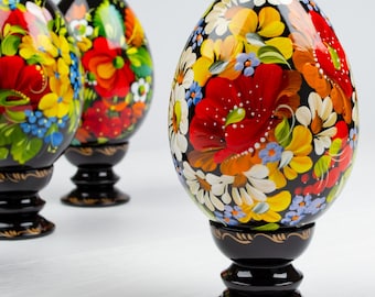 Op de afbeelding: Drie zwarte houten eieren versierd met kleurrijke bloemenpatronen. De eieren staan op zwarte houten standaards.