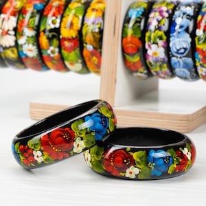 Könnte beinhalten: Zwei schwarze Holzarmreifen mit handbemalten Blumendesigns. Die Armreifen zeigen rote, blaue, weiße und grüne Blumen auf schwarzem Hintergrund.
