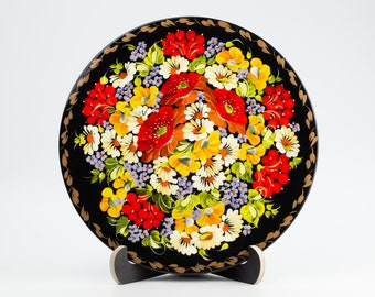 May include: A black round wooden plate with a hand-painted floral design. The plate features red, yellow, white, and green flowers with a black background and gold trim.