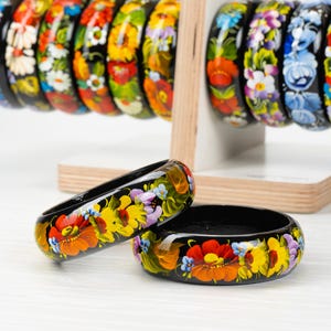 May include: Two black wooden bangles with colorful floral designs painted on them. The bangles are stacked on top of each other and are on a white surface.
