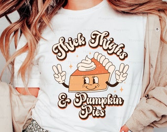 Könnte beinhalten: Weißes T-Shirt mit einem Retro-Grafikdesign, das eine lächelnde Kürbis-Torte mit einem fehlenden Stück zeigt. Der Text "Thick Thighs & Pumpkin Pies" ist in einer fetten, stilisierten Schrift geschrieben.