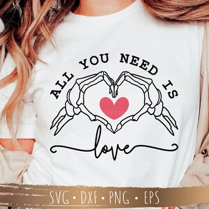 May include: White t-shirt with black text that reads "All you need is love" with a skeleton hand heart graphic and a pink heart in the center.