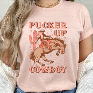 May include: A light pink t-shirt with a graphic of a cowboy riding a bucking bronco, a cactus, and the words "Pucker Up Cowboy" in a vintage font. The design is in shades of brown and pink.
