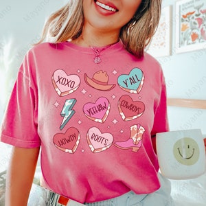 Peut inclure: T-shirt rose avec un motif imprim&eacute; vache, comprenant des c&oelig;urs avec diff&eacute;rentes phrases comme "XOXO", "Y'all", "Yeehaw", "Howdy", "Boots", et "Cowboys".