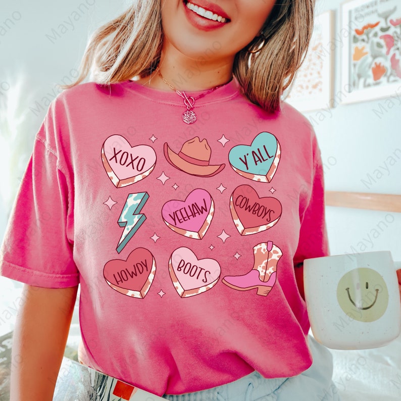 Peut inclure: T-shirt rose avec un motif imprim&eacute; vache, comprenant des c&oelig;urs avec diff&eacute;rentes phrases comme "XOXO", "Y'all", "Yeehaw", "Howdy", "Boots", et "Cowboys".
