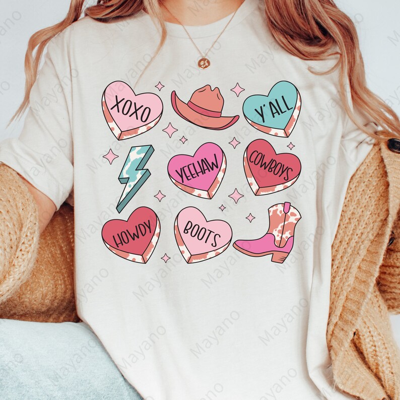 Peut inclure: Un t-shirt blanc avec un motif de c&oelig;ur rose, bleu et rouge. Chaque c&oelig;ur porte un mot diff&eacute;rent, notamment "XOXO", "Howdy", "Yeehaw", "Boots", "Cowboys" et "Y'all".