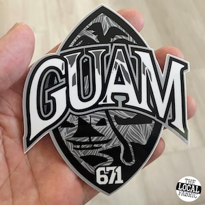 May include: Black and white sticker with the word "GUAM" in a bold, stylized font. The sticker features a palm tree and a boat design with the number "671" at the bottom. The text "THE LOCAL FABRIC" is also visible in the bottom right corner.