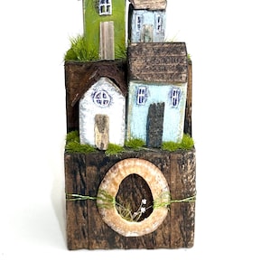 Könnte beinhalten: Eine handgefertigte Holzskulptur mit Miniaturhäusern in Grün-, Blau- und Weißtönen. Die Häuser haben braune Dächer und sind auf einem strukturierten Holzsockel mit einer dekorativen Muschel und Grün angeordnet. Ein einzigartiger Dekorationsartikel.