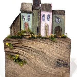 May include: A rustic wooden sculpture featuring a miniature village of four houses with varying colors and architectural details. The houses are mounted on a weathered wooden base with faux greenery and small yellow flowers. The houses have painted details.