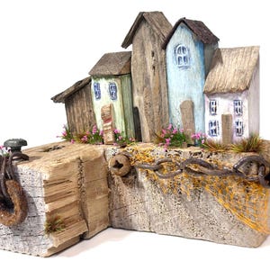 May include: A handcrafted miniature coastal village sculpture. The artwork features several small wooden houses painted in pastel colors, with details like windows and doors. A weathered wooden base includes a rusty metal ring and chain, with decorative flowers and grass.