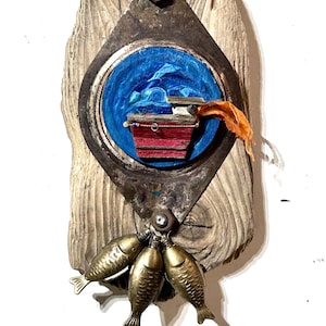 May include: A wall-mounted assemblage art piece featuring a weathered piece of driftwood as a base. A circular metal frame holds a painted blue sea scene with a small red wooden boat. Three metallic gold-toned fish charms hang from the bottom.