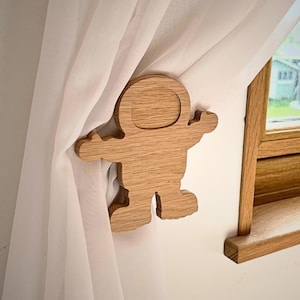 May include: Wooden astronaut curtain tieback. The tieback is shaped like an astronaut with a round helmet and outstretched arms. The tieback is made of light brown wood.
