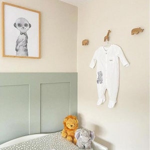 Pu&ograve; includere: Un body bianco per neonato con una stampa di animali grigi &egrave; appeso a un gancio di legno a forma di animale. Il body &egrave; su una parete beige chiaro con un rivestimento verde sottostante. Ci sono tre ganci di legno a forma di animale sul muro, e una stampa incorniciata di un suricato con papillon &egrave; sopra il rivestimento.