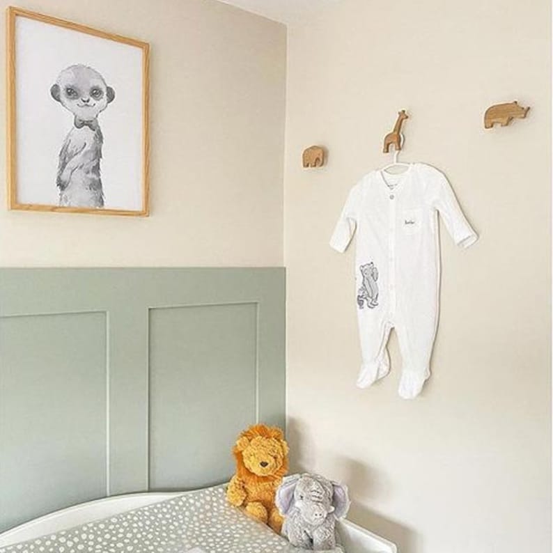 Pu&ograve; includere: Un body bianco per neonato con una stampa di animali grigi &egrave; appeso a un gancio di legno a forma di animale. Il body &egrave; su una parete beige chiaro con un rivestimento verde sottostante. Ci sono tre ganci di legno a forma di animale sul muro, e una stampa incorniciata di un suricato con papillon &egrave; sopra il rivestimento.