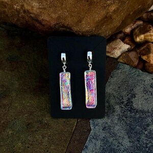 May include: A pair of rectangular dangle earrings with iridescent, colorful glass. The earrings have a silver-colored metal hook and a small silver-colored metal loop connecting the hook to the glass. The glass has a pink, purple, and yellow color scheme.