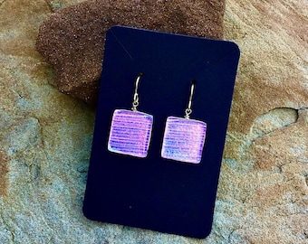 May include: A pair of square, iridescent earrings with silver hooks. The earrings are a light purple color with horizontal lines, displayed on a dark blue card. The background is a textured, tan and brown stone surface.