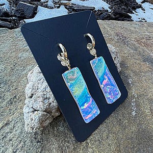 May include: A pair of gold-colored hoop earrings with rectangular, iridescent pendants. The pendants display a mix of blue, green, and pink hues. The earrings are displayed on a black card, set against a natural, rocky background.