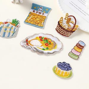 May include: A collection of embroidered food patches, including a bag of crisps, a bread basket, an oyster, a cake, and macarons. The patches are brightly coloured and detailed, with the text "Longancraft" at the top.