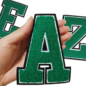 Op de afbeelding: Drie groene viltletters, E, A en Z, met witte randen en een zwarte achtergrond. De letters worden in een hand gehouden.