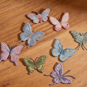 Peut inclure: Plusieurs écussons papillon brodés, aux couleurs variées comme le bleu, le rose, le vert et le violet, sont disposés sur une surface en bois. Les écussons présentent des coutures détaillées et sont de tailles différentes. Le texte "Longancraft" est visible.