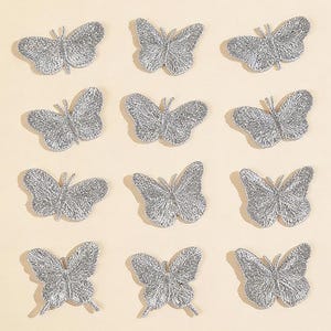 May include: Nine silver glitter butterfly embellishments arranged on a light beige background. The butterflies have detailed wing patterns and are evenly spaced. The text "Longancraft" is displayed at the top of the image.