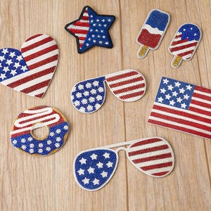 Op de afbeelding: Een verzameling geborduurde patches met een patriottisch thema, waaronder een hart, een ster, twee ijsjes, twee zonnebrillen, een donut en een rechthoekige vlag, allemaal versierd met rood-wit-blauwe sterren- en strepenpatronen.