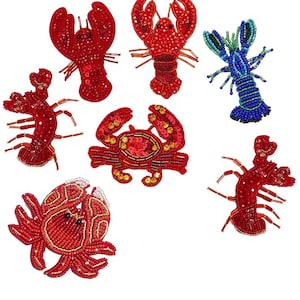 Peut inclure: Une collection d'ornements de crustacés perlés, comprenant des homards et des crabes rouges, et un homard bleu. Les ornements sont ornés de paillettes et de perles. Le texte "Longancraft" est affiché en haut.