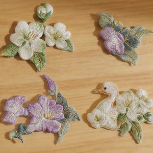 May include: Four embroidered fabric patches featuring floral and animal motifs. The designs include white blossoms with green leaves, purple flowers with foliage, and a white swan surrounded by white flowers, all with detailed thread work.