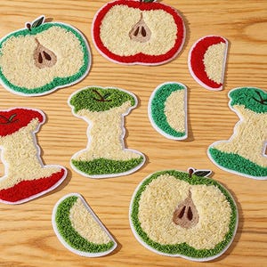 Może przedstawiać: Haftowane naszywki z jabłkami w różnych fazach jedzenia, w kolorach czerwonym, zielonym i kremowym. Naszywki zawierają u góry napis "Longancraft". Naszywki znajdują się na drewnianej powierzchni.