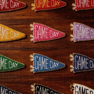 May include: Assortment of colorful felt pennants with the text "GAME DAY" embroidered in white. Each pennant has a gold fringe and is displayed on a dark wood surface. The brand name "Longancraft" is at the top.