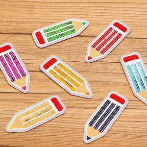 Peut inclure: Plusieurs écussons en forme de crayon de couleur rouge, vert, jaune, rose et bleu. Chaque écusson a une gomme rouge et un bord argenté. Les écussons sont disposés sur une surface en bois. Le texte "Longancraft" est visible.