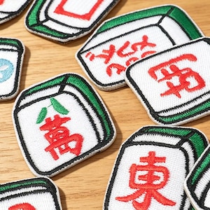 May include: A collection of embroidered patches shaped like mahjong tiles, featuring white surfaces with black borders and green accents. Each patch displays distinct red Chinese characters or symbols, including the East wind and various numeric tiles.