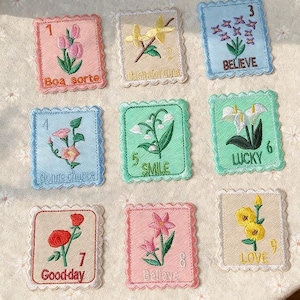 May include: Nine embroidered patches with floral designs and positive affirmations. Each patch features a different flower, number, and phrase like "Believe" or "Good-day". The patches have scalloped edges and are in various pastel colours.