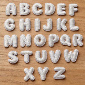 May include: A set of white embroidered alphabet letter patches featuring a rounded, puffy font style. Each letter from A to Z is individually stitched with a light blue border. The text Longancraft is displayed at the top with two pink hearts.