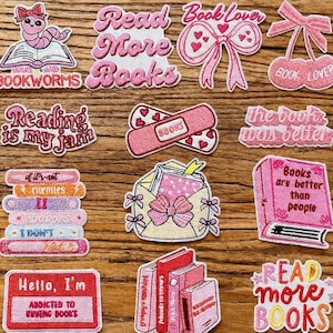 Op de afbeelding: Een verzameling geborduurde patches met een boeken thema. De patches tonen zinnen als "Read More Books" en "Book Lover". Kleuren zijn roze, rood en wit. Sommige patches tonen boeken, strikken en een boekenwurm.