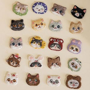 Può includere: Toppe ricamate a forma di gatto in vari colori e design, tra cui nero, grigio, arancione e bianco. Le toppe presentano diverse espressioni di gatti e sono disposte su una superficie beige chiaro. La parola "Longancraft" è in alto.