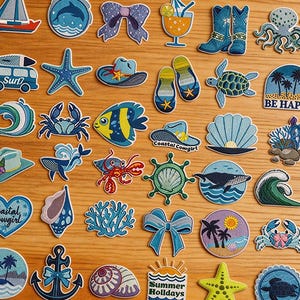 Può includere: Una collezione di toppe ricamate a tema costiero ed estivo. Presentano vari motivi come una barca a vela, una stella marina, un granchio, un pesce e la scritta "Summer Holidays". Le toppe sono blu, gialle e bianche. Altri disegni includono un fiocco, stivali da cowboy e un cocktail.