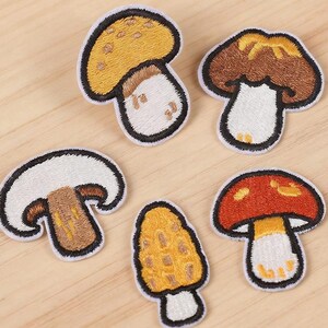 Può includere: Cinque toppe ricamate a forma di fungo di varie forme e colori, con cappelli gialli, marroni, bianchi e rossi su gambi bianchi, il tutto bordato con filo nero. Il testo Longancraft è visibile in alto con piccole icone a forma di cuore.