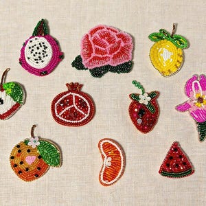 Op de afbeelding: Een verzameling van kralen broches met fruit en bloemen, waaronder een drakenvrucht, roos, citroen, appel, granaatappel, aardbei, sinaasappelschijf en watermeloen. De broches zijn gemaakt van kleurrijke kralen. De tekst "Longancraft" staat bovenaan.