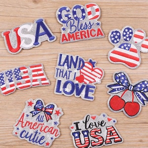 May include: A selection of embroidered patches with patriotic themes. Designs include the text "USA", "God Bless America", "Land That I Love", and "I Love USA". Other patches feature a paw print, cherries, and a bow with stars and stripes.