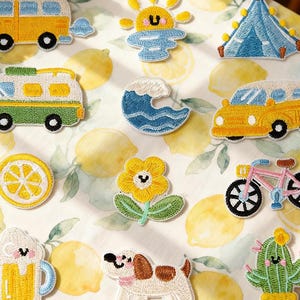 Może przedstawiać: Haftowane naszywki z różnymi wzorami, w tym żółtym kamperem, uśmiechniętym słońcem, niebieskim namiotem, falą, żółtym samochodem, plasterkiem cytryny, kwiatem, rowerem, kuflem piwa, psem i kaktusem. Naszywki znajdują się na tle z wzorem cytryny. U góry znajduje się napis "Longancraft".