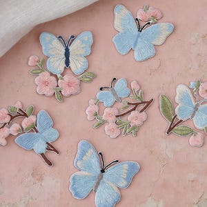 May include: Six embroidered patches featuring light blue butterflies and pink cherry blossom flowers with green stems and leaves. The text Longancraft is displayed at the top, accompanied by two small pink hearts and a pair of small white wings on the sides.