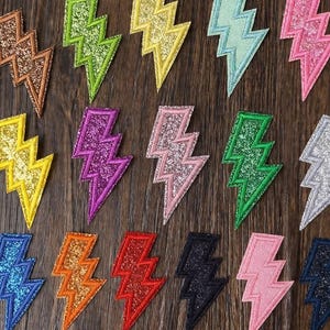 May include: A selection of colourful, glitter-covered lightning bolt patches. The patches, in shades like gold, green, and pink, are set against a wooden background. Each bolt has a contrasting border. The text "Longancraft" is visible.