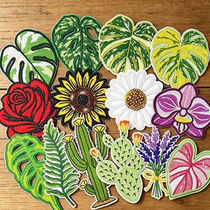 Op de afbeelding: Een verzameling geborduurde patches met diverse botanische ontwerpen, waaronder monstera-bladeren, een rode roos, een gele zonnebloem, een witte margriet, een paarse orchidee, cactussen, lavendel en een blad met roze nerven.