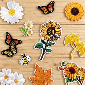 May include: A collection of embroidered fabric patches featuring various designs including white daisies, orange monarch butterflies, yellow sunflowers, a bee, a maple leaf, and ginkgo leaves. The text Longancraft is visible at the top.