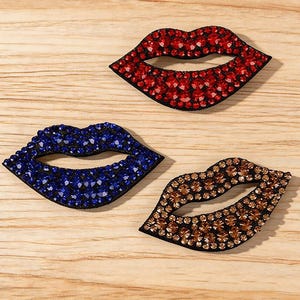 Op de afbeelding: Drie met strassteentjes bezette lipvormige patches in rood, blauw en brons. De patches zijn gerangschikt op een licht oppervlak met houtnerf. Het woord "Longancraft" wordt bovenaan de afbeelding weergegeven.